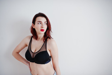 Red haired girl with a big bust on black bra and jeans against white wall at empty room.