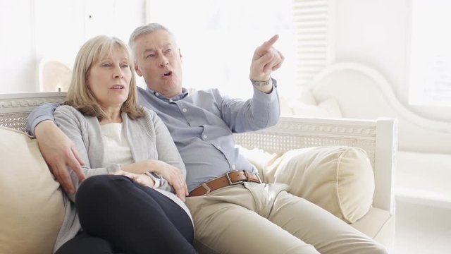 Mature couple watching tv as the husband points out something he doesn't like 
