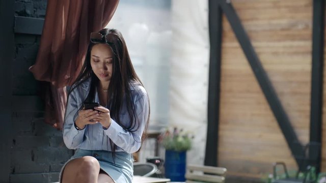 Attractive young Asian woman sitting at outdoor terrace in summer cafe and texting on smartphone