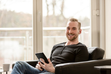 Smiling blonde man sitting indoors in his living room holding a digital tablet looking at camera with a big smile on his face.