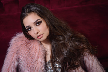 Beauty vogue style close up portrait. Pretty sexy brunette model girl wearing silver party dress and coat posing at red sofa background in room of the hotel. Wavy long hair bright make up smoky eyes