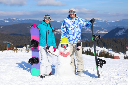 Happy couple with snowman and snowboards at ski resort. Winter vacation