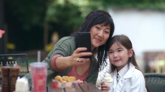 Asian mother and cute little girl with ice cream sitting together at cafe table outdoors and taking selfie with smartphone