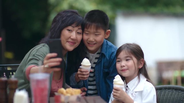Cute little Asian boy and girl holding ice cream and smiling at smartphone camera while taking selfie with mother at cafe table outdoors