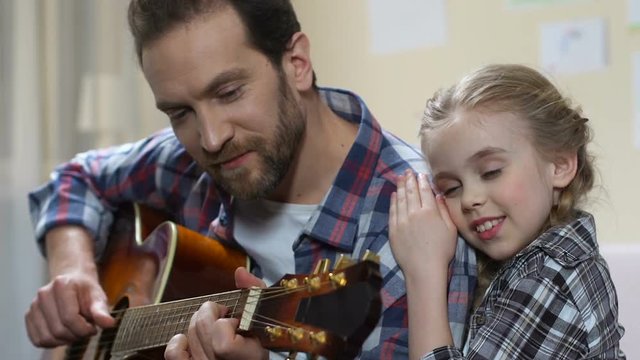 Little girl hugging and listening father playing guitar, creative home leisure