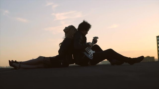couple in rock-n-roll style siting on the rooftop with flapping hairs on the wind at sunset sky background, man playing on guitar