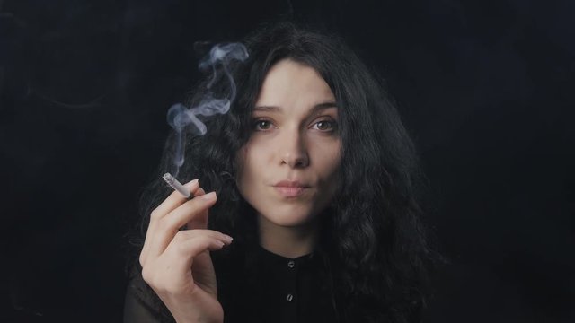 Portrait of young brunette woman with curly hair is smoking a cigarette and looking at the camera on the dark background