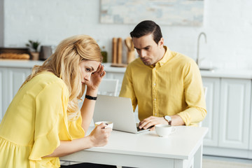 Upset woman with coffee sitting by man working on laptop in kitchen