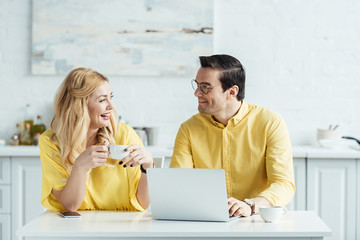 Pretty couple with coffee looking at each other and sitting by laptop on kitchen table
