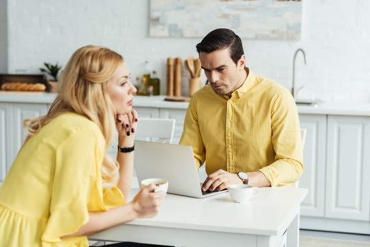 Sad girlfriend holding coffee and sitting by her boyfriend working on laptop in kitchen