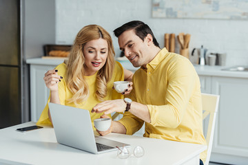 Man and woman drinking coffee and looking at laptop screen by kitchen table