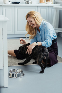 Smiling Blonde Woman Playing With Frenchie Dog On Kitchen Floor