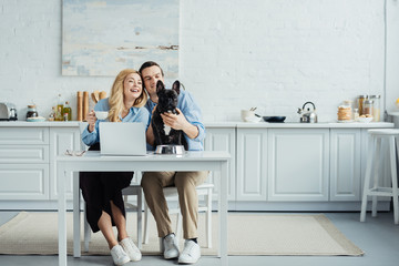 Smiling couple with coffee sitting by table with laptop in kitchen and stroking Frenchie dog