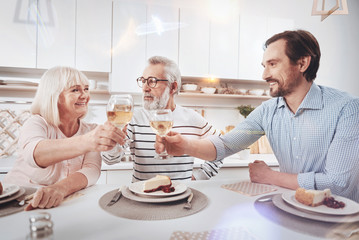 Happy to see each other. Pleasant delighted adult man giving a toast and sitting at the table while enjoying time with his loving parent
