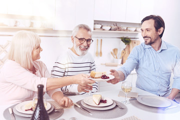 Thank you. Cheerful delighted mother givign piece of cheesecake her adult son while sitting at the table together and enjoyign meal