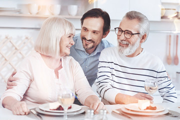 Warm relationships. Cheerful delighted aged couple sitting at the table while his adult son embracing them and resting in the kitchen