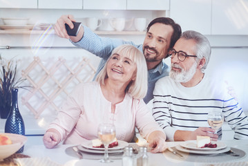 Just smile. Positive delighted adult son takign selfies with his aged parents while sitting together in the kitchen and expressing gladness
