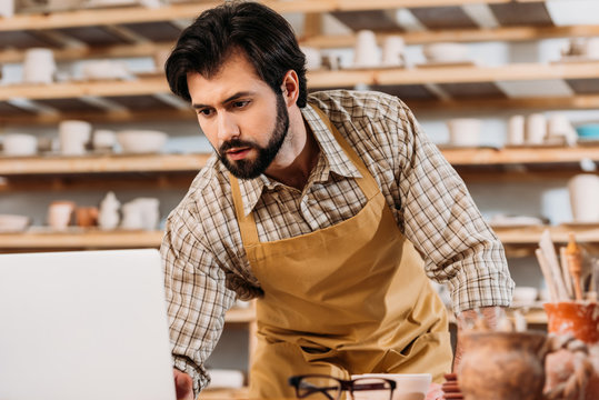 male potter in apron working with laptop in workshop