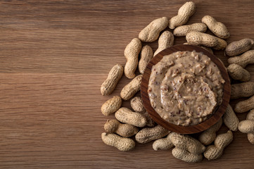 American breakfast, sweet food and snack high in calories concept with a bowl of crunchy peanut butter surrounded by peanuts still in shell, isolated on wood background with copy space