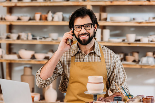 smiling male potter talking on smartphone in workshop