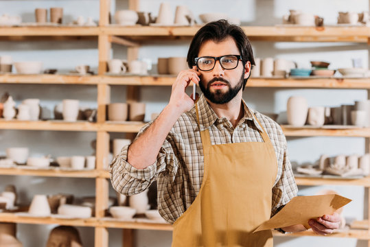 male potter holding envelope and talking on smartphone in workshop