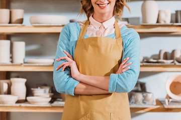 cropped view of potter with crossed arms standing near shelves with ceramic dishware