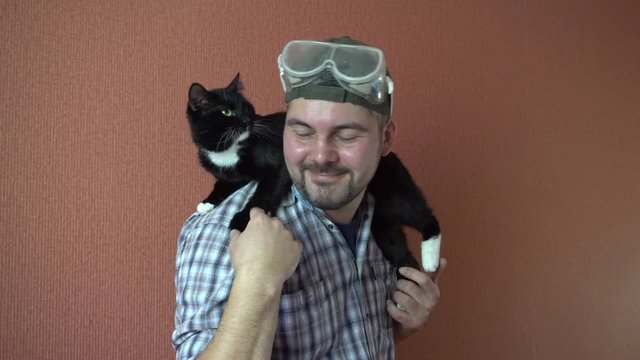 A man holds on his shoulders the dark the cat. Fluffy pet. Stroking the fur with your hand. In a plaid shirt. The green cap. Safety glasses on the head. 4K video