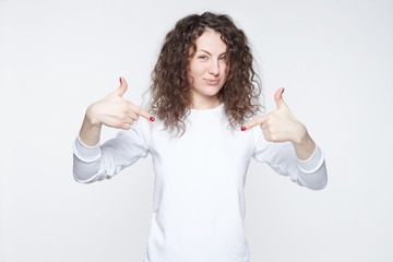Horizontal shot of cheerful female with happy look points at white casual t shirt with copy space...