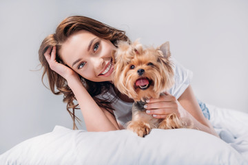 portrait of young smiling woman with yorkshire terrier resting on bed isolated on grey