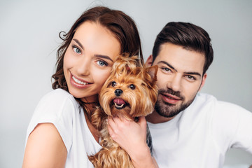 portrait of young smiling couple with yorkshire terrier looking at camera isolated on grey