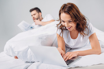 Obraz premium selective focus of smiling woman using laptop while lying on bed together with boyfriend isolated on grey