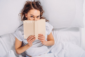 obscured view of woman covering face with book and looking at camera while lying in bed