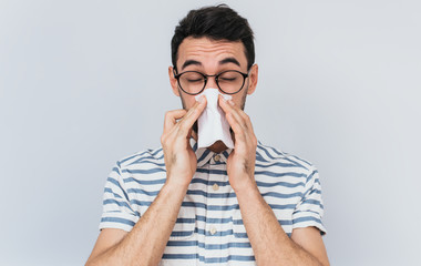 Horizontal portrait of unhealthy handsome man wearing striped shirt and glasses, blowing nose into tissue. Male have flu, virus or allergy against white background. Healthy medicine and people concept
