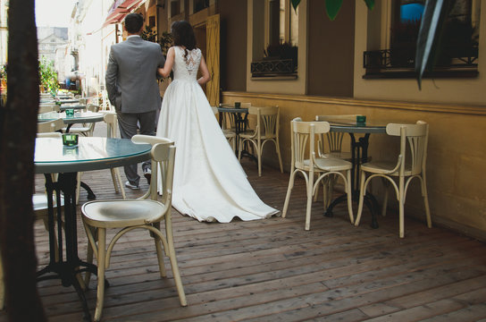 Elegant wedding couple is walking in old ancient town street cafe. Slim bride in satin dress with train. Groom in grey checkered suit. Wooden floor and summer atmosphere.