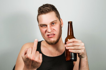 Frowning young gyu showing middle finger and holding bottle on beer, on gray background.