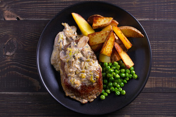 Beef steak diane with mushroom and leek cream sauce, potato fries and green peas in black plate on wooden background. overhead