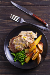 Beef steak diane with mushroom and leek cream sauce, potato fries and green peas in black plate on wooden background. overhead