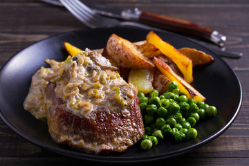 Beef steak diane with mushroom and leek cream sauce, potato fries and green peas in black plate on wooden background