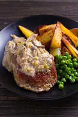 Beef steak diane with mushroom and leek cream sauce, potato fries and green peas in black plate on wooden background