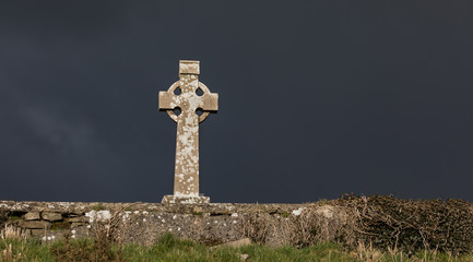 Celtic cross gravestone behind soon wall of a cemetery in rural Ireland, dark stormy rain clouds and sunlight