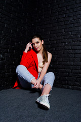 Stylish brunette girl on red jacket against studio black brick wall.