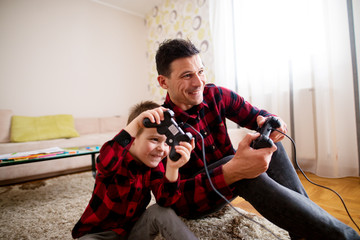 Young cheerful excited father and son in the same red shirt playing console games with gamepads...