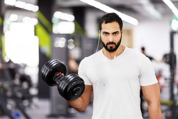Strong sexy bearded man wears white t-shirt and headphones does arm dumbbells workout in the gym