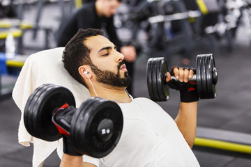Fototapeta na wymiar Bearded man wears white t-shirt and headphones does pushup workout with dumbbells on the branch in the gym