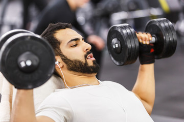 Bearded man wears white t-shirt and headphones does pushup workout with dumbbells on the branch in the gym