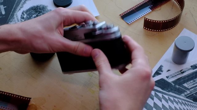 Man loading 35mm film into vintage film camera with black and white photos and developed film laying around the wooden desk
