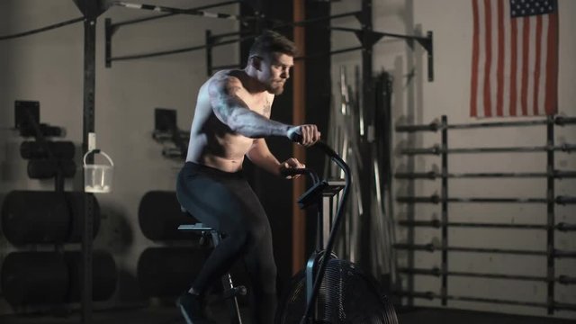 Determined shirtless athlete with tattooed body working out in gym and riding bicycle exerciser.