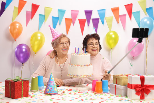 Two cheerful elderly women with party hats and a birthday cake taking a selfie
