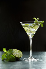 Summer fresh cocktail with lime slices, crushed ice and mint leaves in a martini glass backlit on black background
