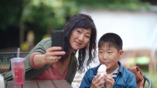 Tracking shot Asian woman smiling at camera of smartphone and taking picture with little son while he eating ice cream in outdoor cafe